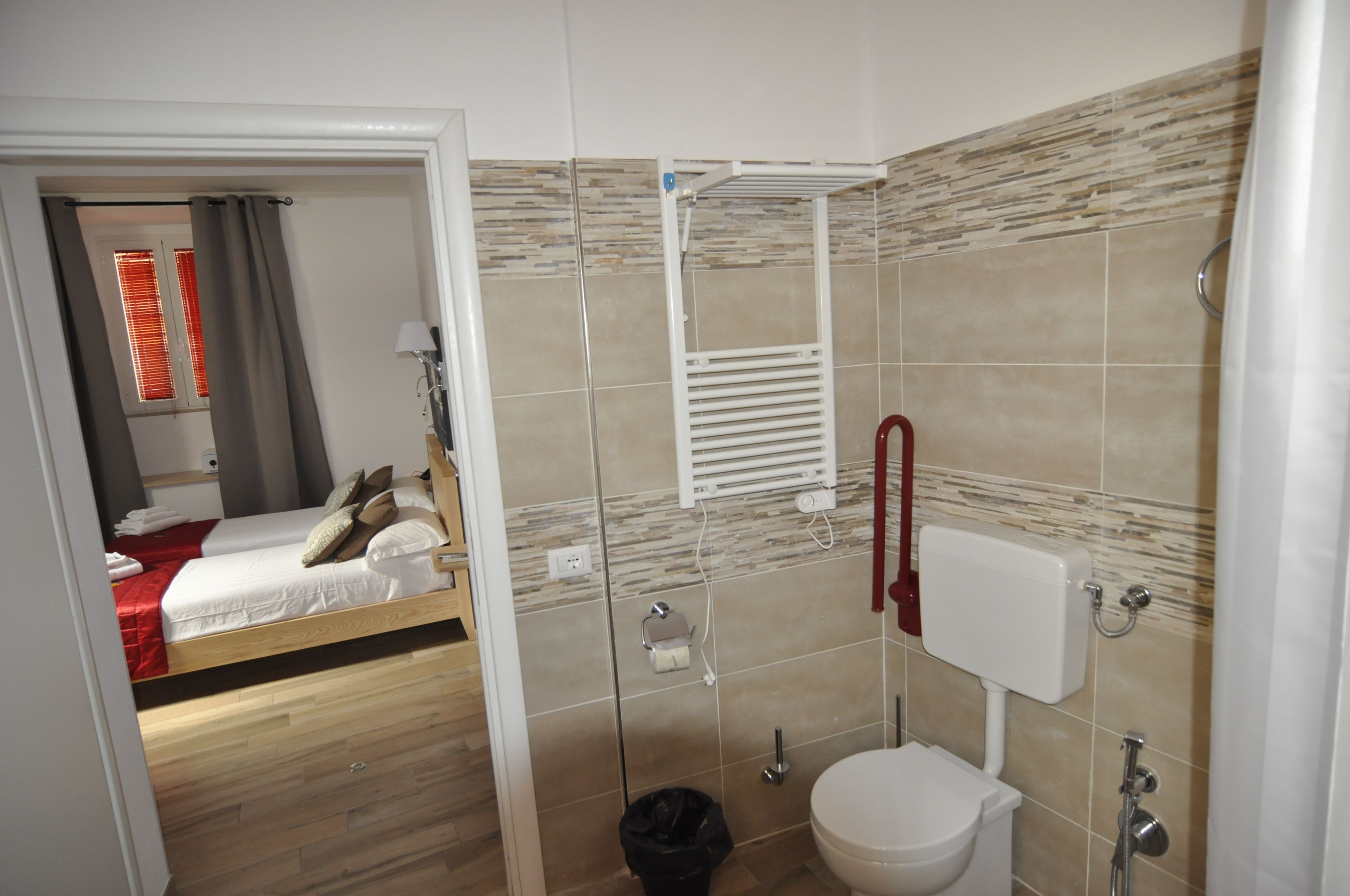 Een kleine hotelkamer met een houten vloer, een bed met rode beddengoed, een douche met een glazen deur en een toilet met een witte tank.