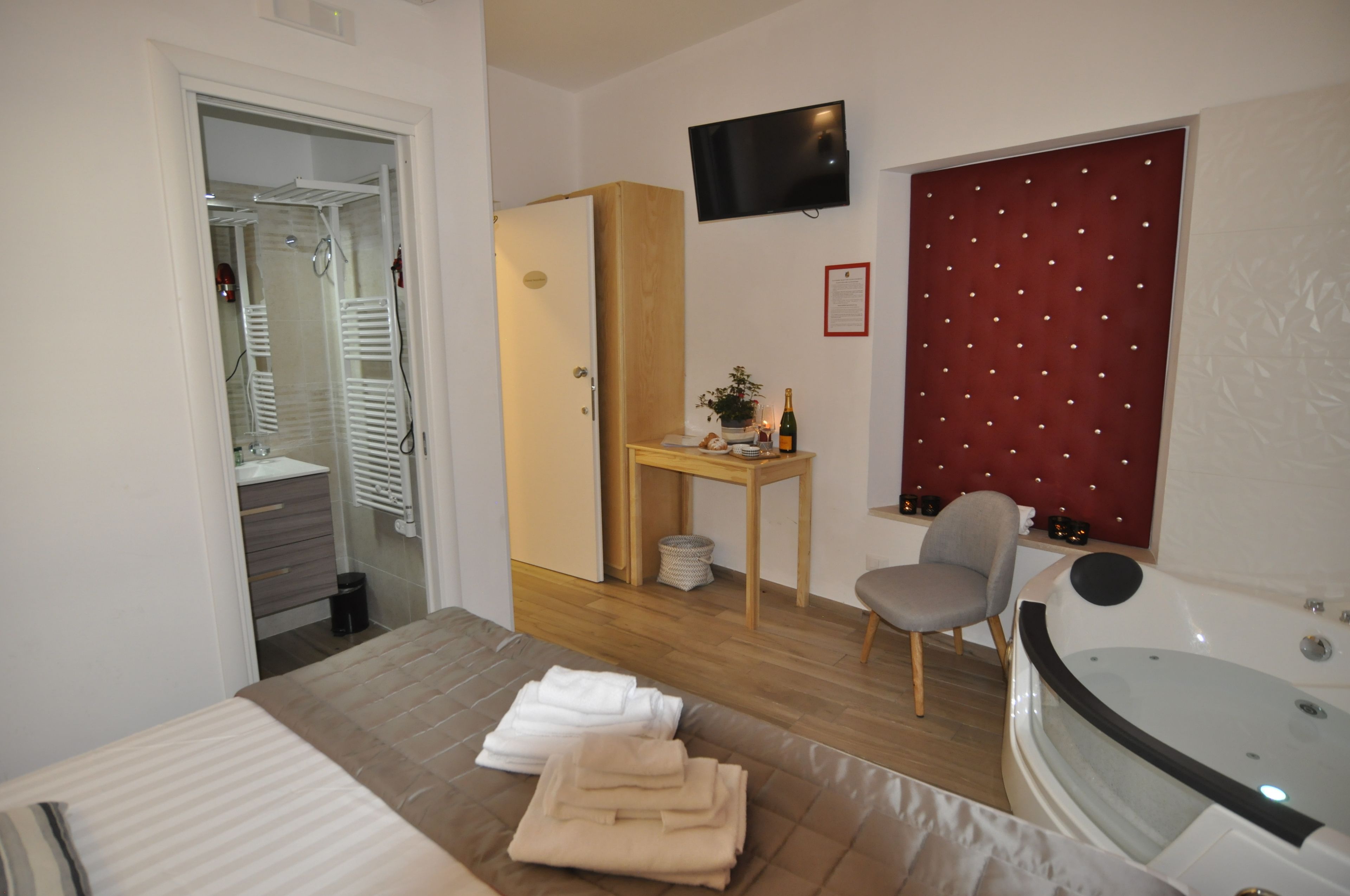 A hotel room with a bed, a desk, a chair, a TV, a red headboard, a bathtub, and a closet with a drying rack.