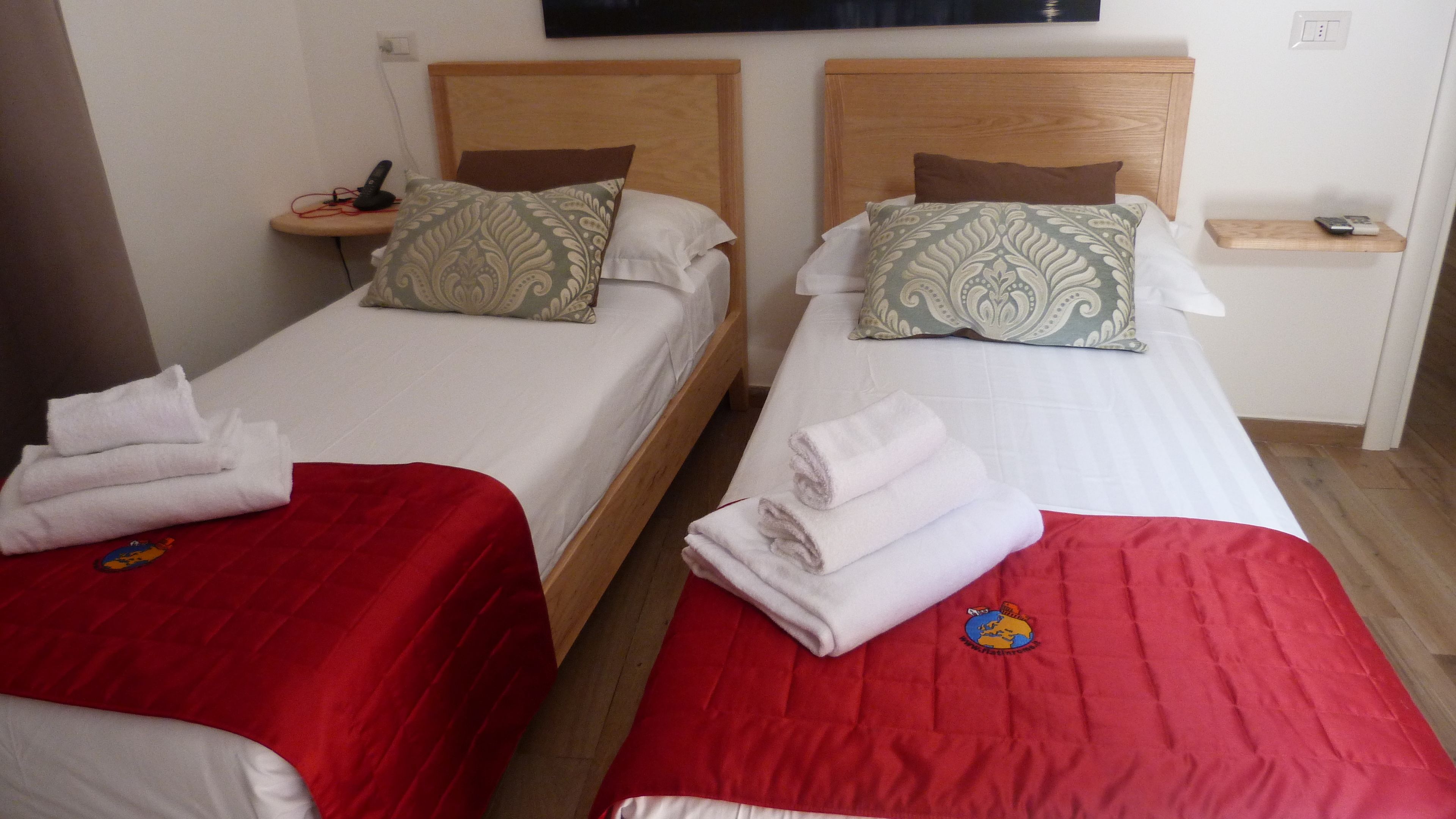 Two single beds with red and white bedding, a wooden headboard, a bedside table with a phone and remote, and a small shelf with a remote.