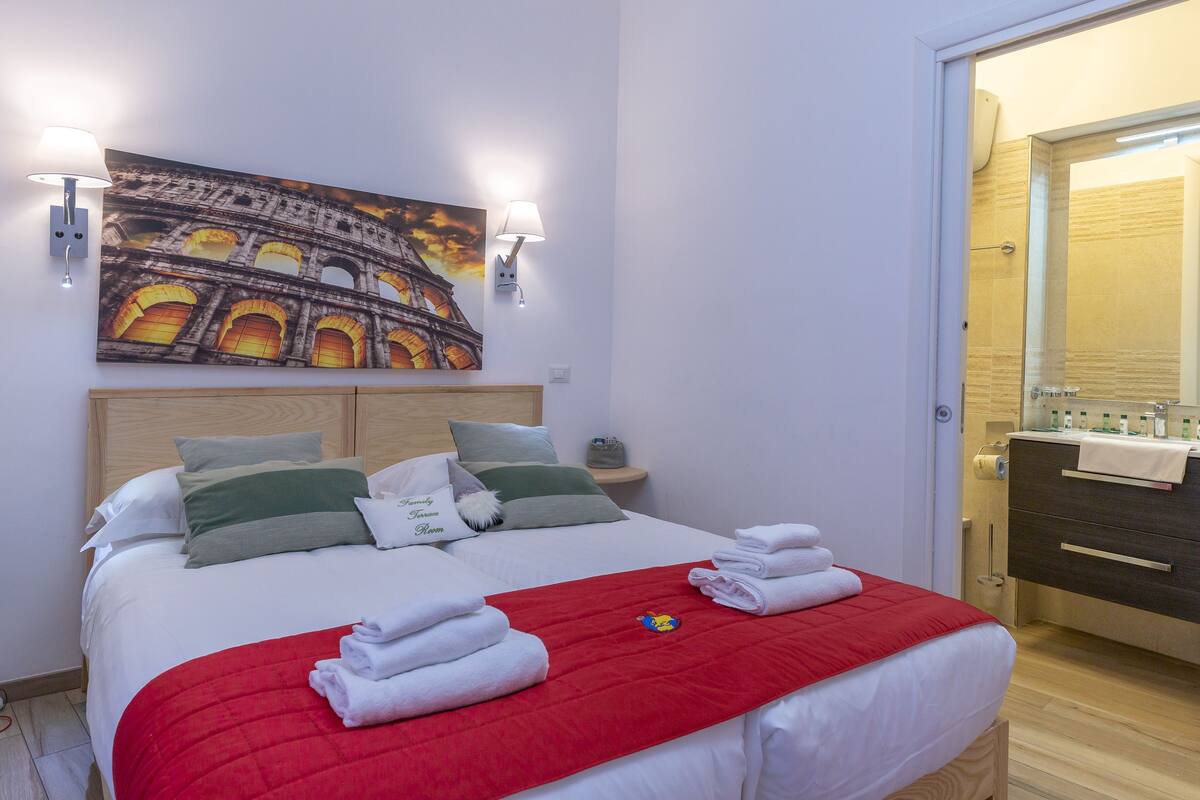 Une chambre d’hôtel avec un lit, une couverture rouge, des serviettes blanches, un tableau représentant le Colisée, et une salle de bain avec un miroir et des articles de toilette.