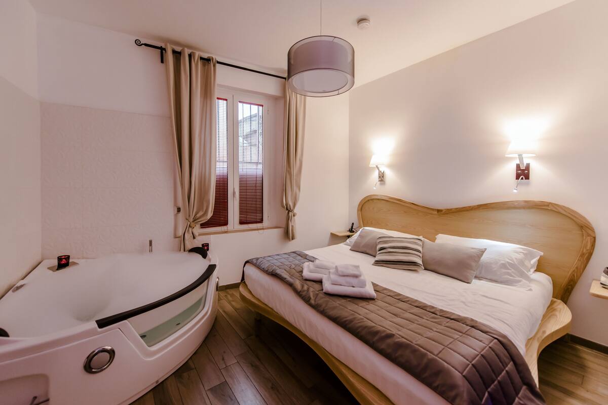 Une chambre d’hôtel avec un lit, une tête de lit en bois, une lampe de chevet, une fenêtre avec des rideaux et une baignoire.