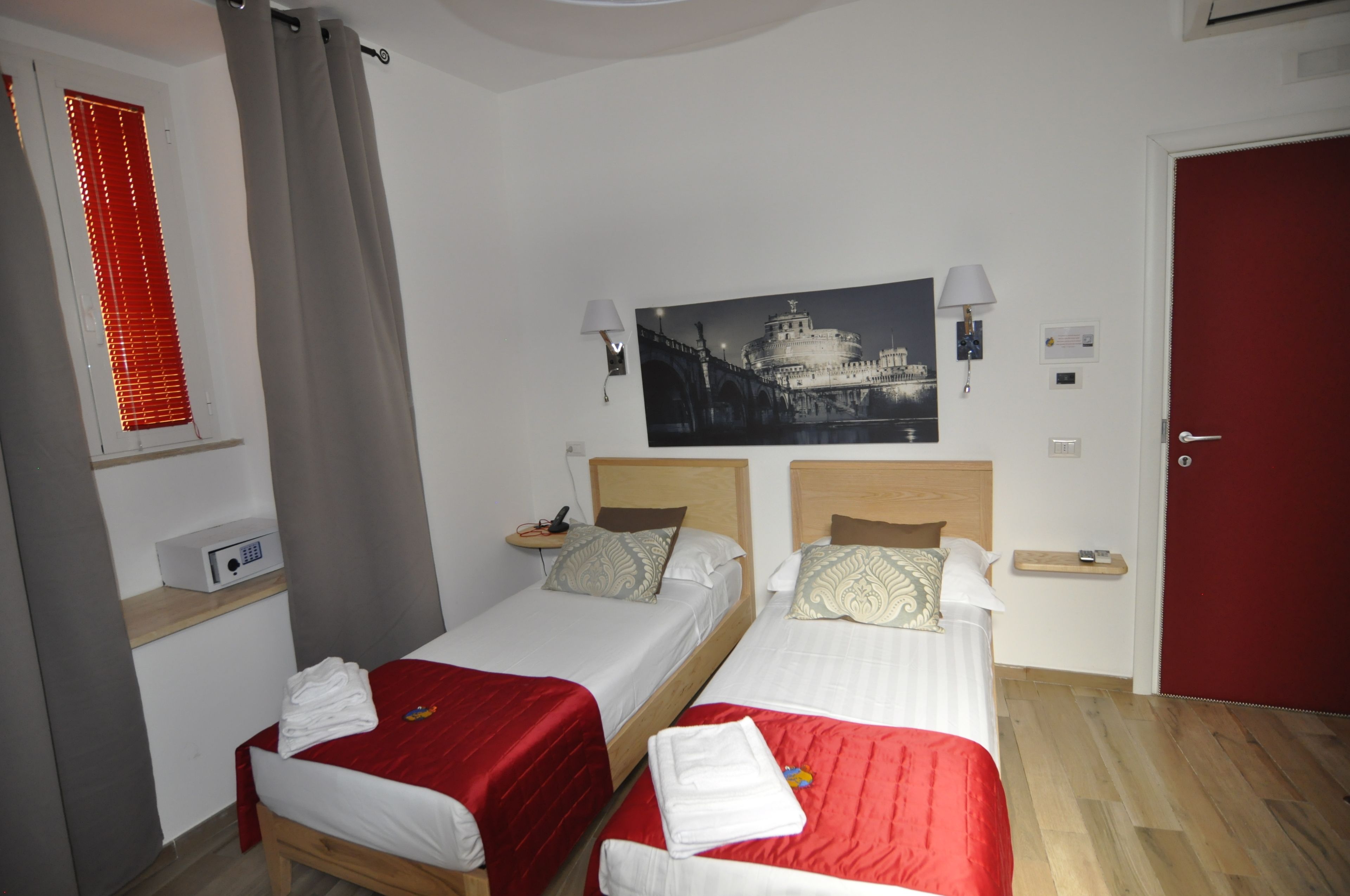 A hotel room with two single beds, a red door, a wall-mounted painting, bedside lamps, and a small bedside table with a lamp.