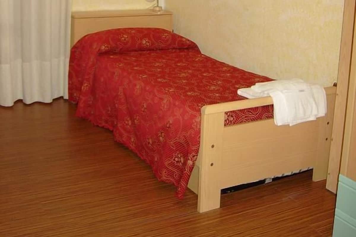Une petite chambre à coucher, bien rangée, avec un lit simple, une table de chevet, un tableau encadré au mur et une fenêtre avec des rideaux.
