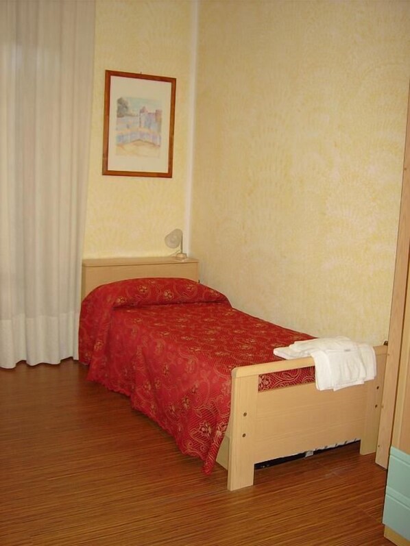 Un dormitorio pequeño y ordenado con una cama individual, una mesita de noche, un cuadro enmarcado en la pared y una ventana con cortinas.