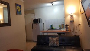 A compact living space with a kitchenette, a sofa with a patterned cushion, and a wall-mounted light fixture. - APARTAHOTEL DIARIO CONCEPCIÓN (Concepción)