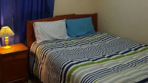 A bedroom with a bed, a nightstand with a lamp, a window with blue curtains, and a ceiling light. - APARTAHOTEL DIARIO CONCEPCIÓN (Concepción)