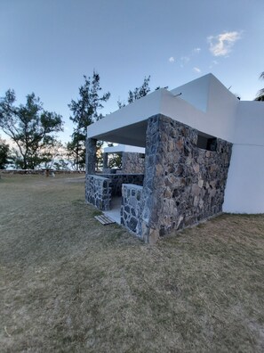 Estructura moderna al aire libre con muros de piedra y techo blanco, rodeada de césped y árboles.