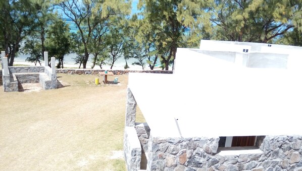 Edificio de paredes de piedra con fachada blanca, patio con césped y árboles al fondo.