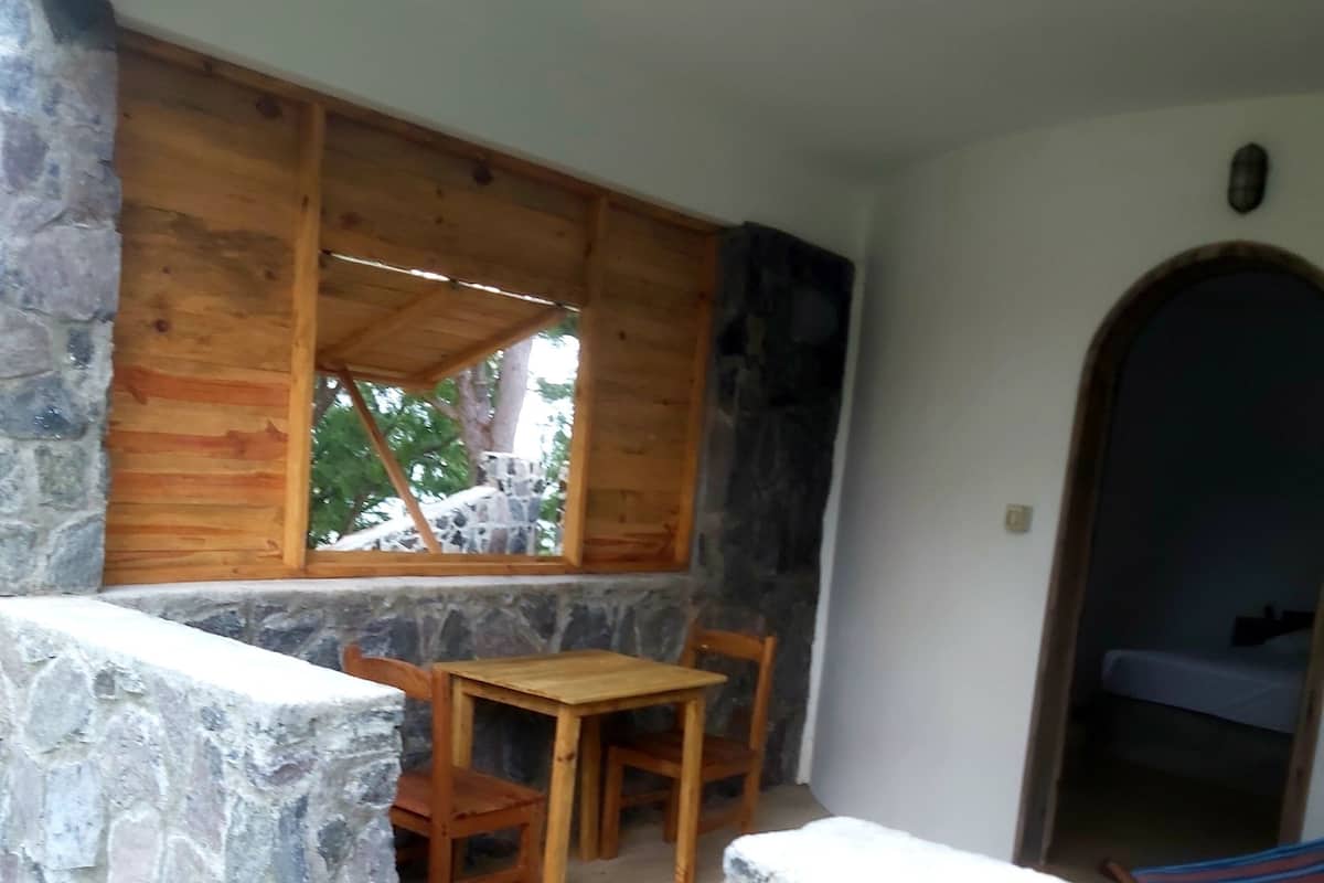 A stone-walled patio with a wooden table and chairs, a window overlooking greenery, and a stone wall leading to a door.