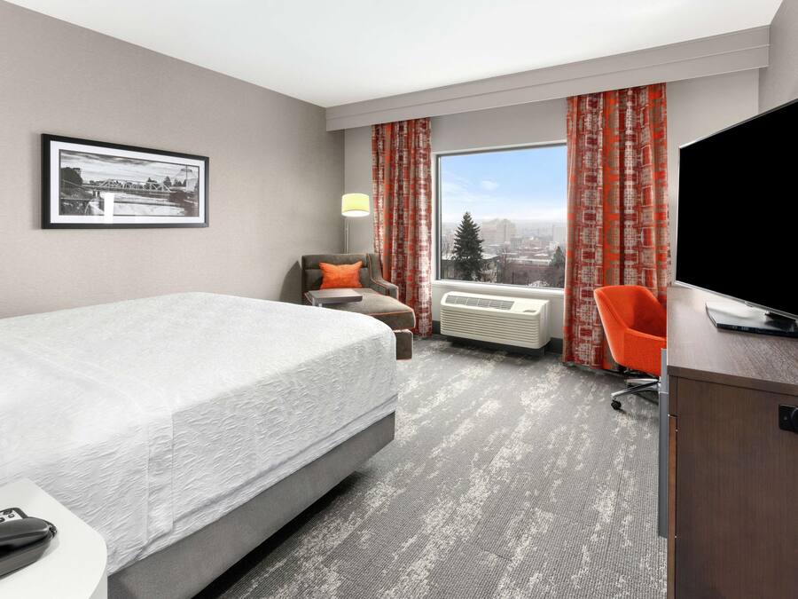 Hotelkamer met een bed, bureau met televisie, stoel, raam met gordijnen, airconditioning en een ingelijst schilderij aan de muur.