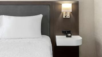 Una cama bien hecha con un almohadón blanco y un cabecero, una mesita de noche empotrada con un lavamanos blanco y un reloj digital, y una lámpara de pared.