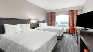 A hotel room with two beds, a desk, a TV, and a view of the city.