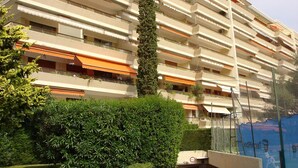 Edifício residencial com vários andares, sacadas, um arbusto em primeiro plano e uma obra em construção ao fundo.