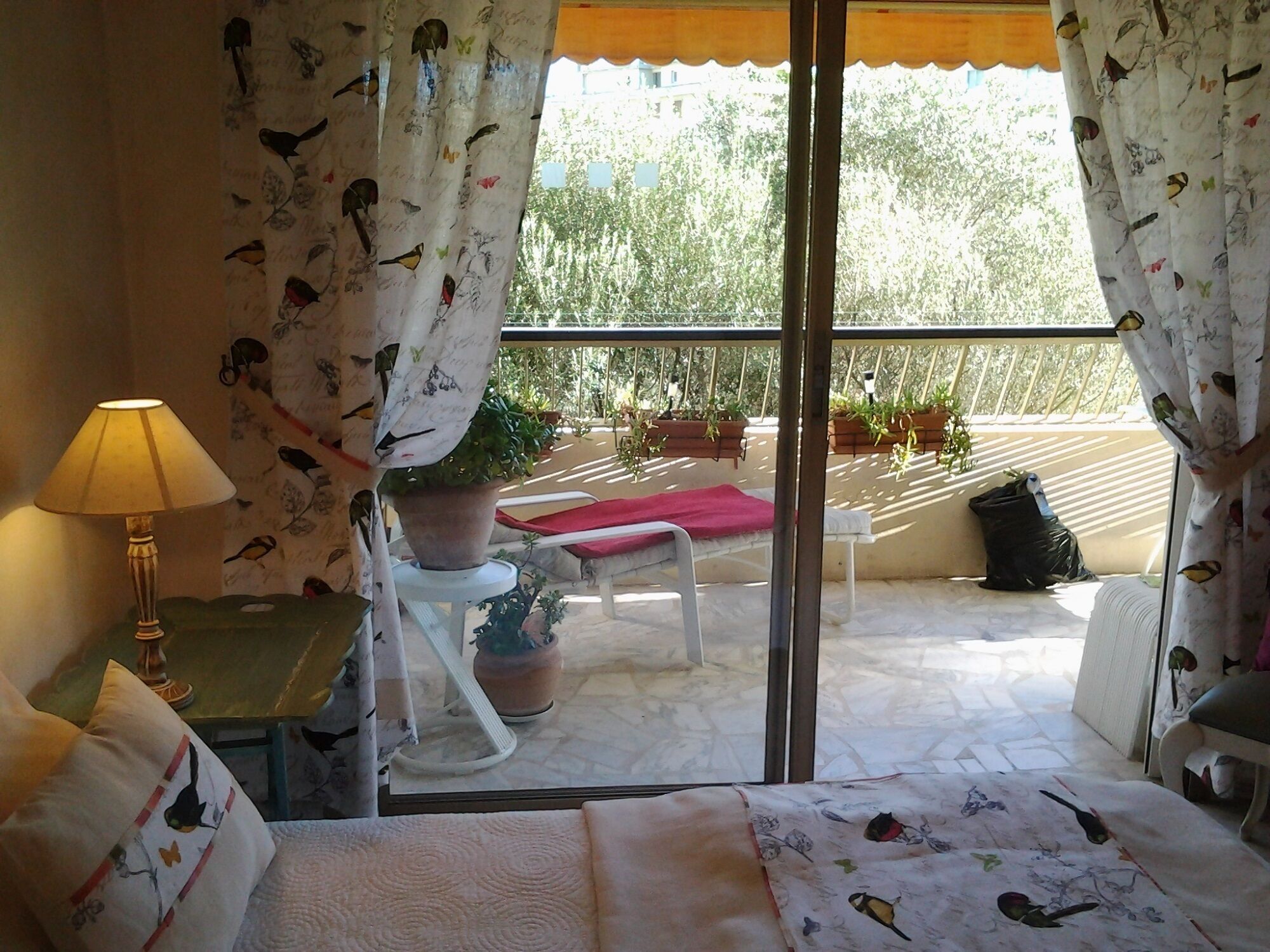A room with a bed, a table with a lamp, a chair, a balcony with potted plants, and a view of trees.