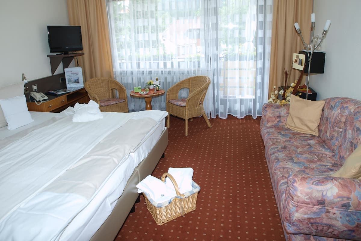 A hotel room with a bed, a sofa, two chairs, a small table with a vase, and a television.
