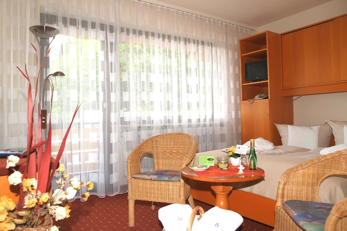A hotel room with a bed, two wicker chairs, a small table with a bottle and glass, and a large window with sheer curtains.