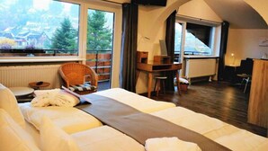 A cozy room with a bed, a desk, and a chair. There are large windows with a view of trees and buildings. - Ferienhotel Schwarzwälder Hof (Feldberg)