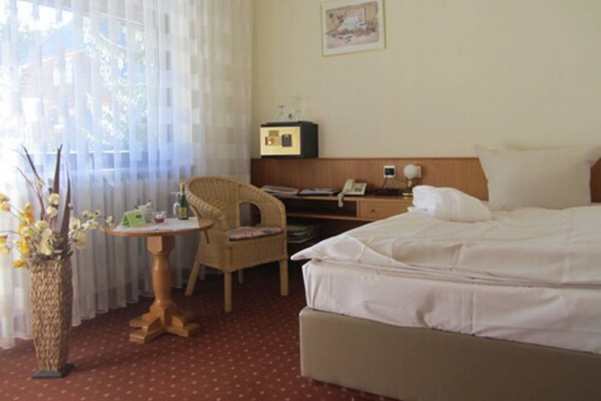 A hotel room with a bed, a desk, a chair, a small table, and a window with sheer curtains.