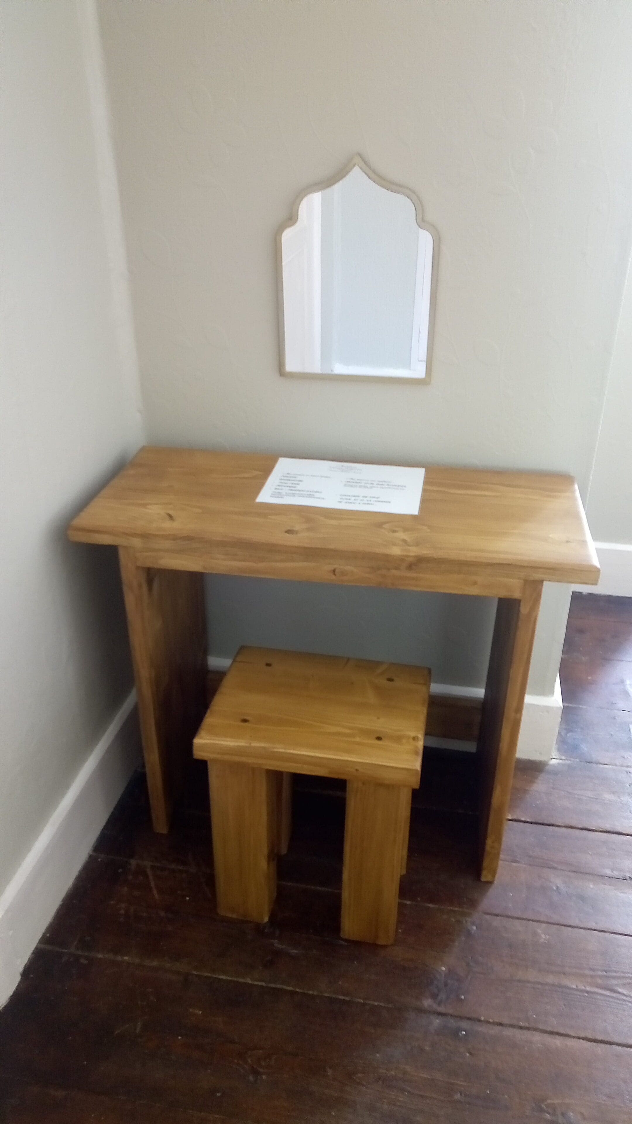 A wooden desk with a matching bench, a wall-mounted mirror, and a piece of paper on the desk.
