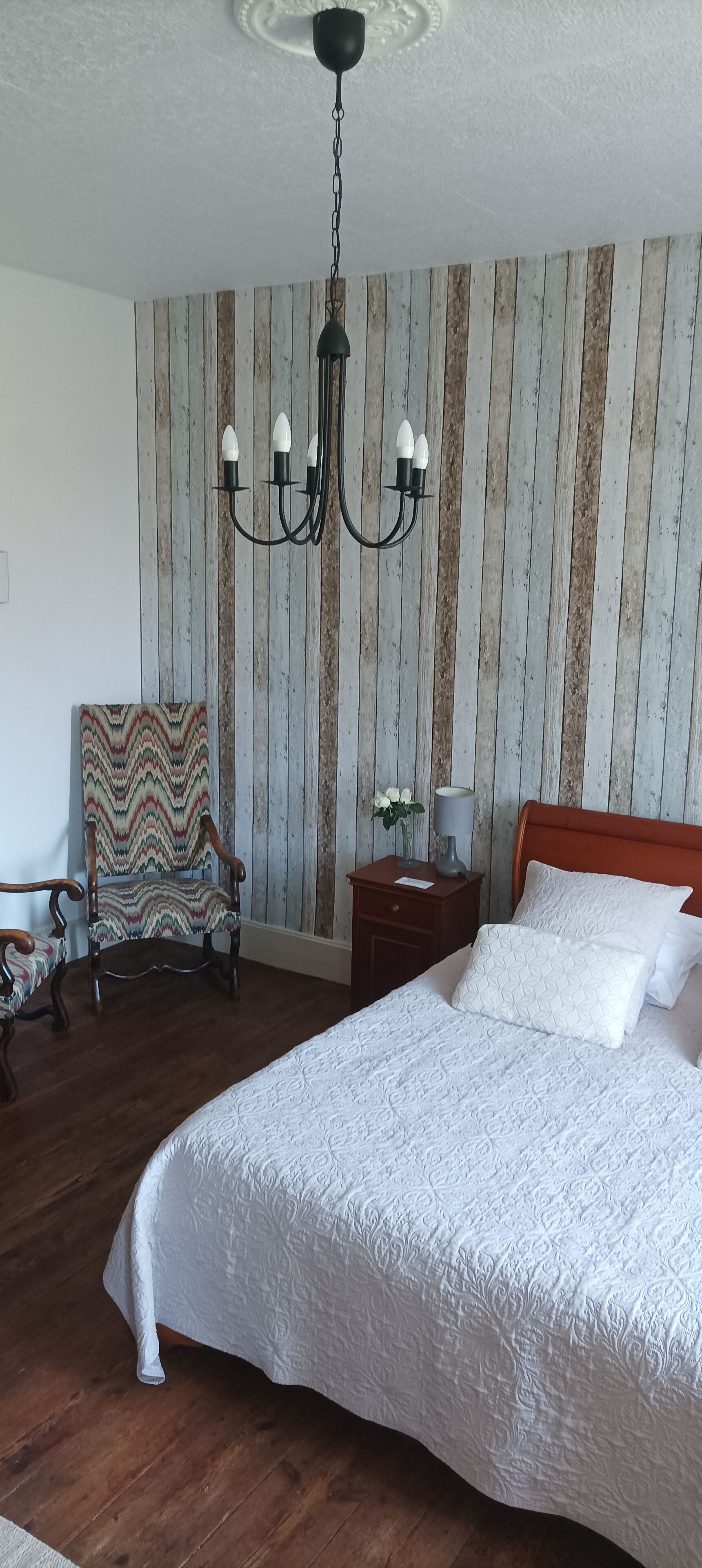 Une chambre à coucher avec un grand lit, une chaise en bois, une table de chevet et un lustre.