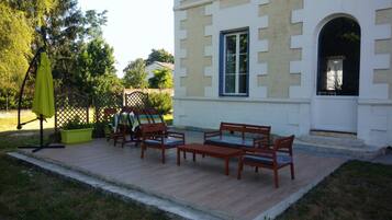 Eine Terrasse im Freien mit Holzmöbeln, einem grünen Sonnenschirm und einem weißen Gebäude mit einem Fenster und einem Bogenportal.