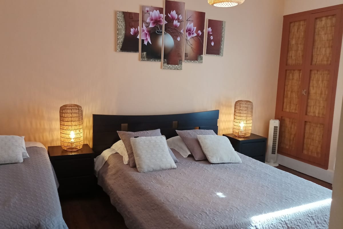 A bedroom with two beds, nightstands with lamps, a wall-mounted artwork, and a wooden door.