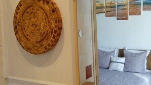 Un baño con un adorno de pared de madera, un espejo que refleja una cama con almohadas y un estante con artículos de tocador.