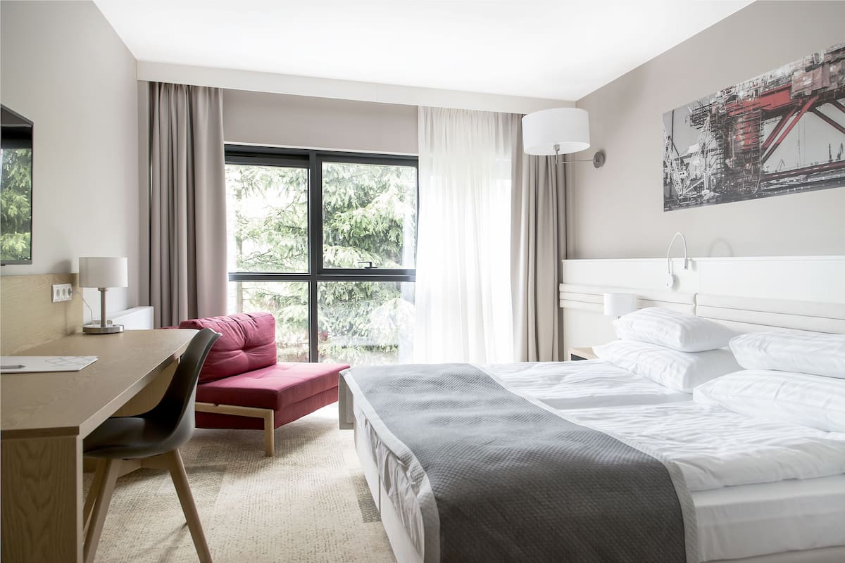 A hotel room with a bed, a desk, a chair, a red sofa, and a window with curtains.
