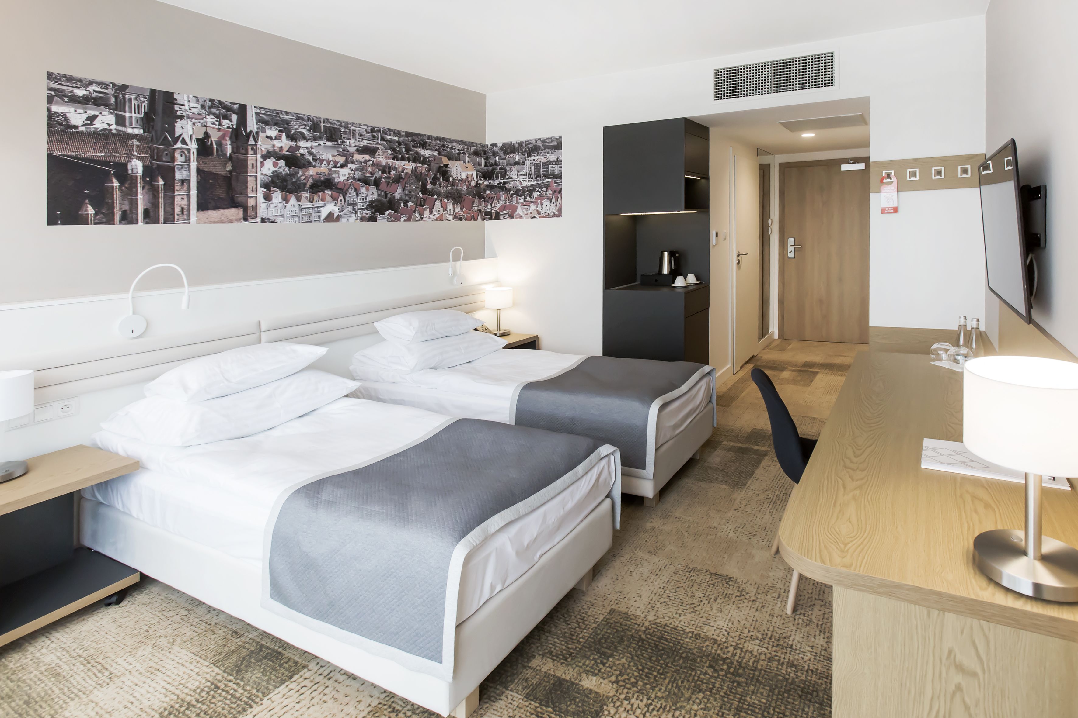 A hotel room with two beds, a desk, a TV, and a wall with a cityscape mural.