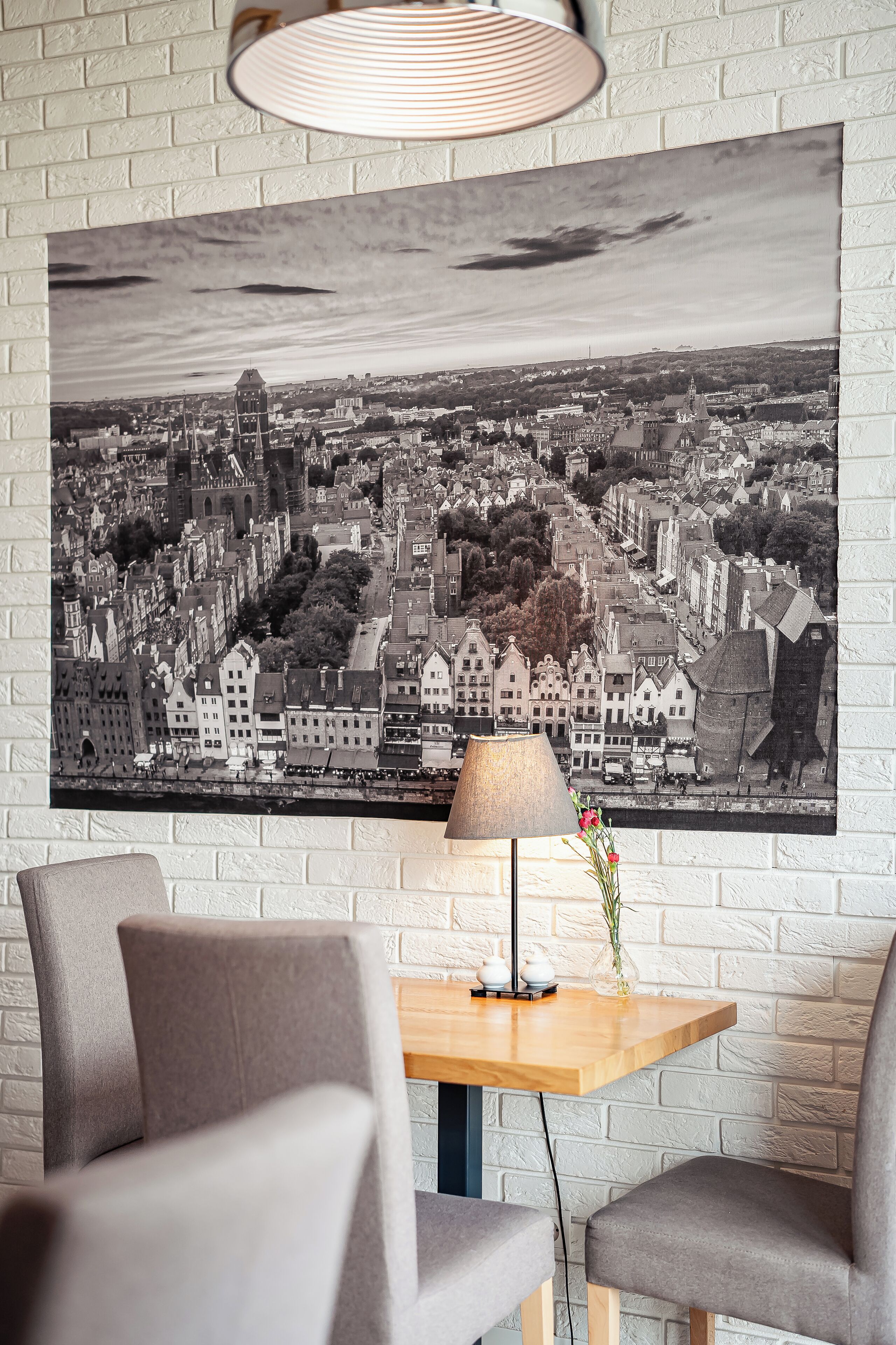 Una habitación con una fotografía grande en blanco y negro de un paisaje urbano, una mesa de madera con una lámpara y flores, y dos sillas grises.