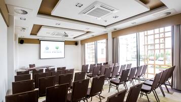 Un salón de conferencias con filas de sillas, una pantalla de proyector y ventanas amplias.