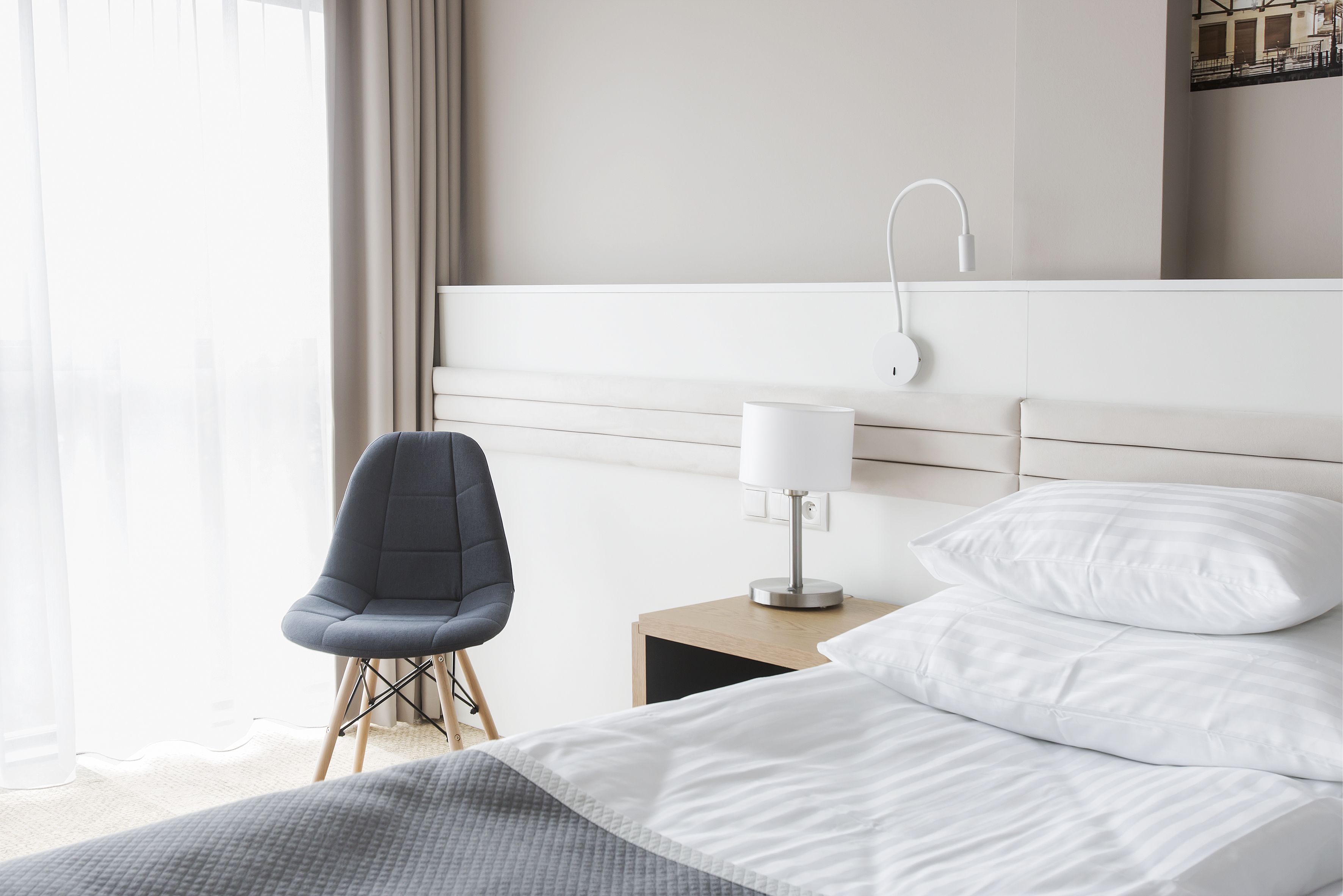 A hotel room with a bed, a chair, a nightstand with a lamp, and a view of the outdoors.