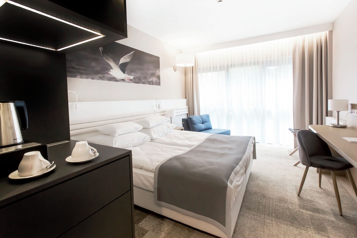 A modern hotel room with a neatly made bed, a desk with a chair, a large window with curtains, and a painting on the wall.