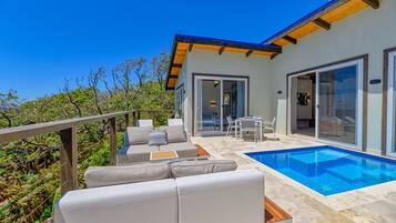 A modern outdoor pool area with a wooden deck, a seating area with cushions, and a glass door leading to an interior space.
