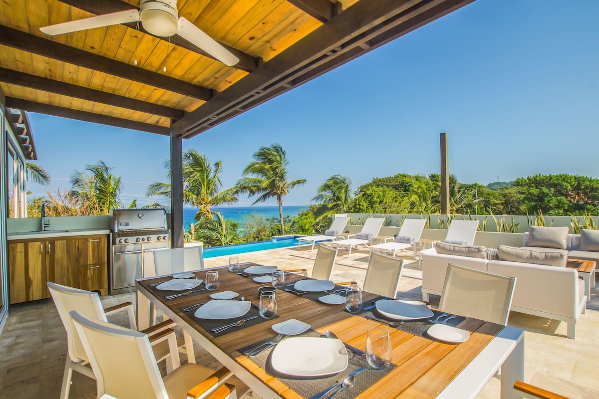 Un área de comedor al aire libre con una mesa de madera para seis personas, rodeada de sillas blancas, con vista a una piscina y palmeras.