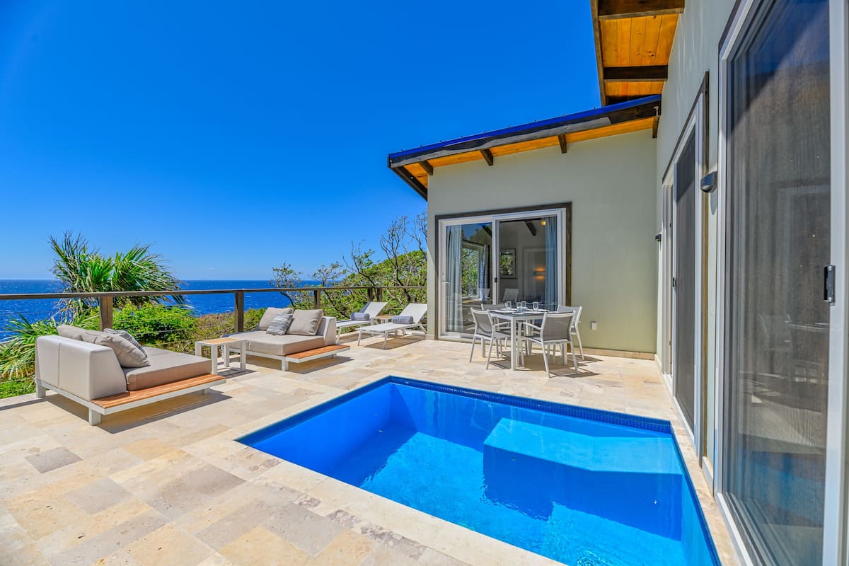 Un área de piscina moderna al aire libre con vista al mar, equipada con sillones de descanso y una mesa de comedor.