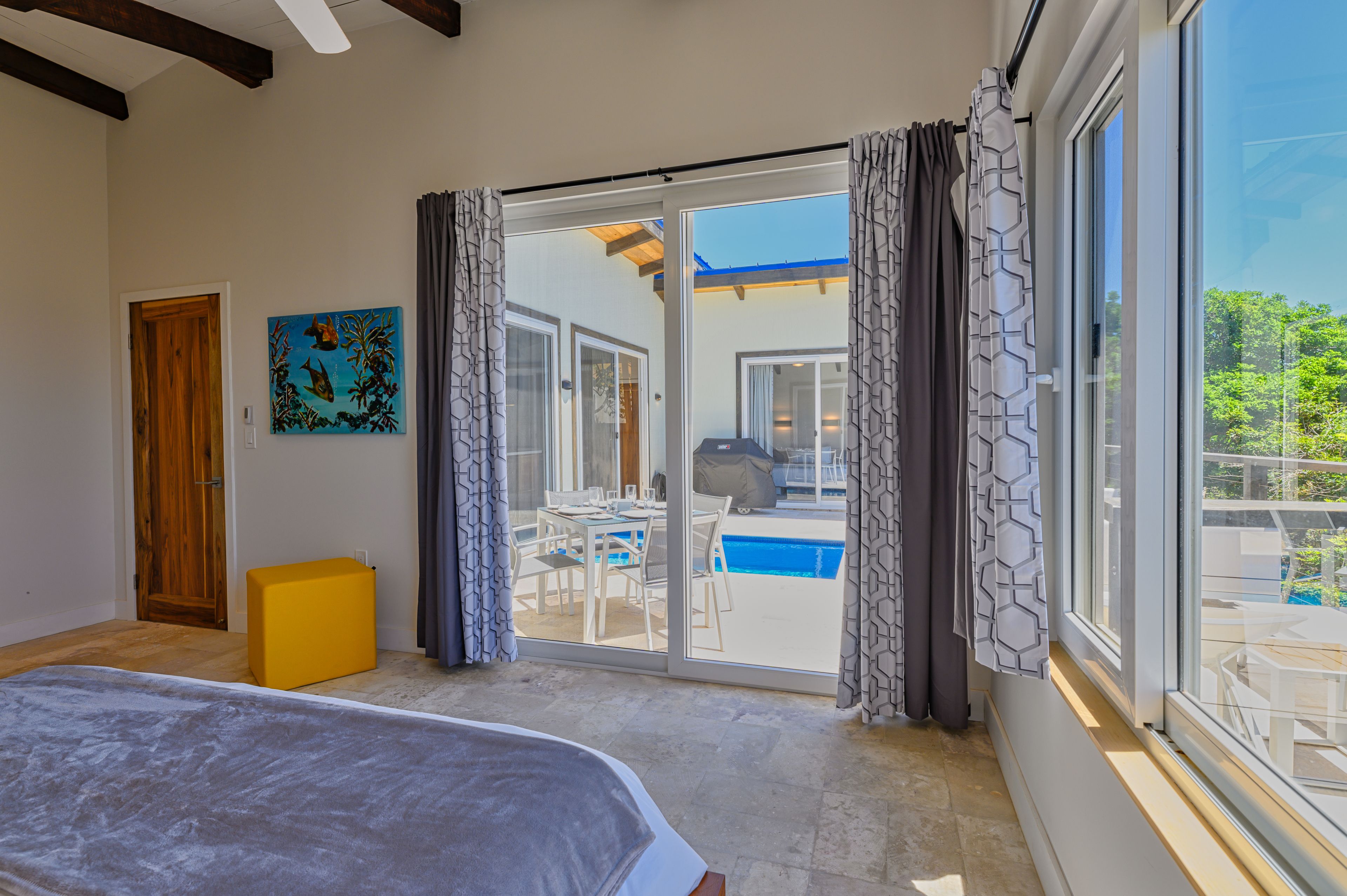 A bedroom with a bed, a wooden door, a yellow ottoman, a painting, and a sliding glass door leading to a balcony with a pool and outdoor furniture.