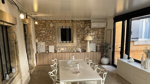 Uma sala de jantar com parede de pedra, uma mesa comprida, cadeiras brancas e uma janela com vista.