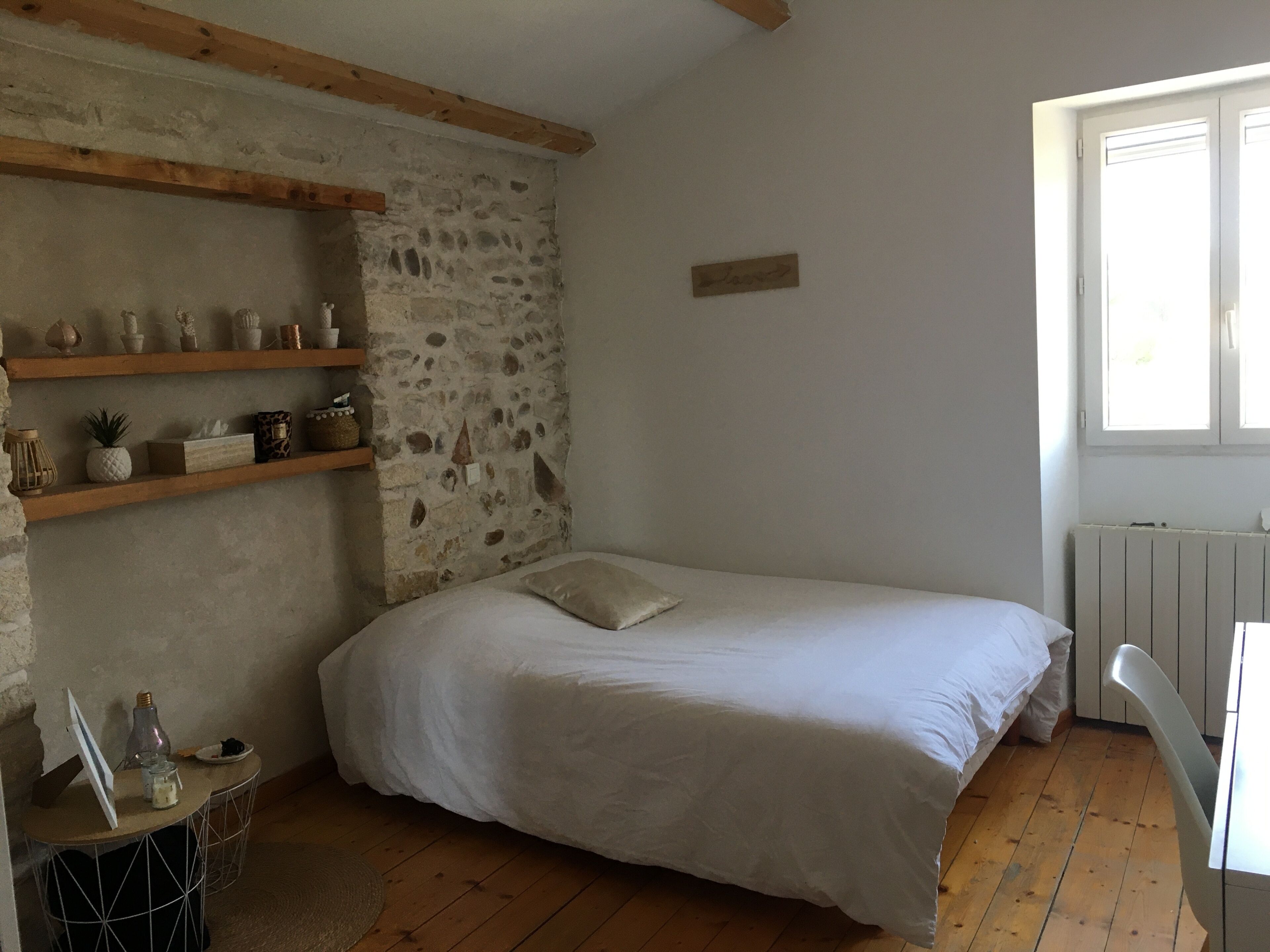 Une chambre avec un lit, des étagères en bois, un mur en pierre, une petite table et une fenêtre.