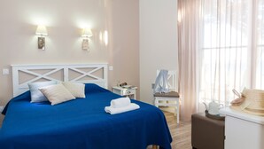 Una cama bien tendida con cubrecama azul y almohadas blancas, una mesita de noche con una lámpara y una ventana con cortinas translúcidas.