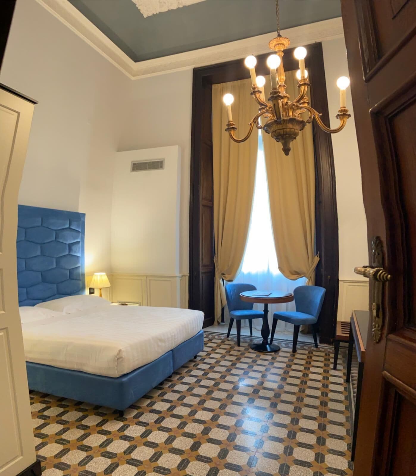 A hotel room with a large bed, a chandelier, a small table with two chairs, and a patterned floor.