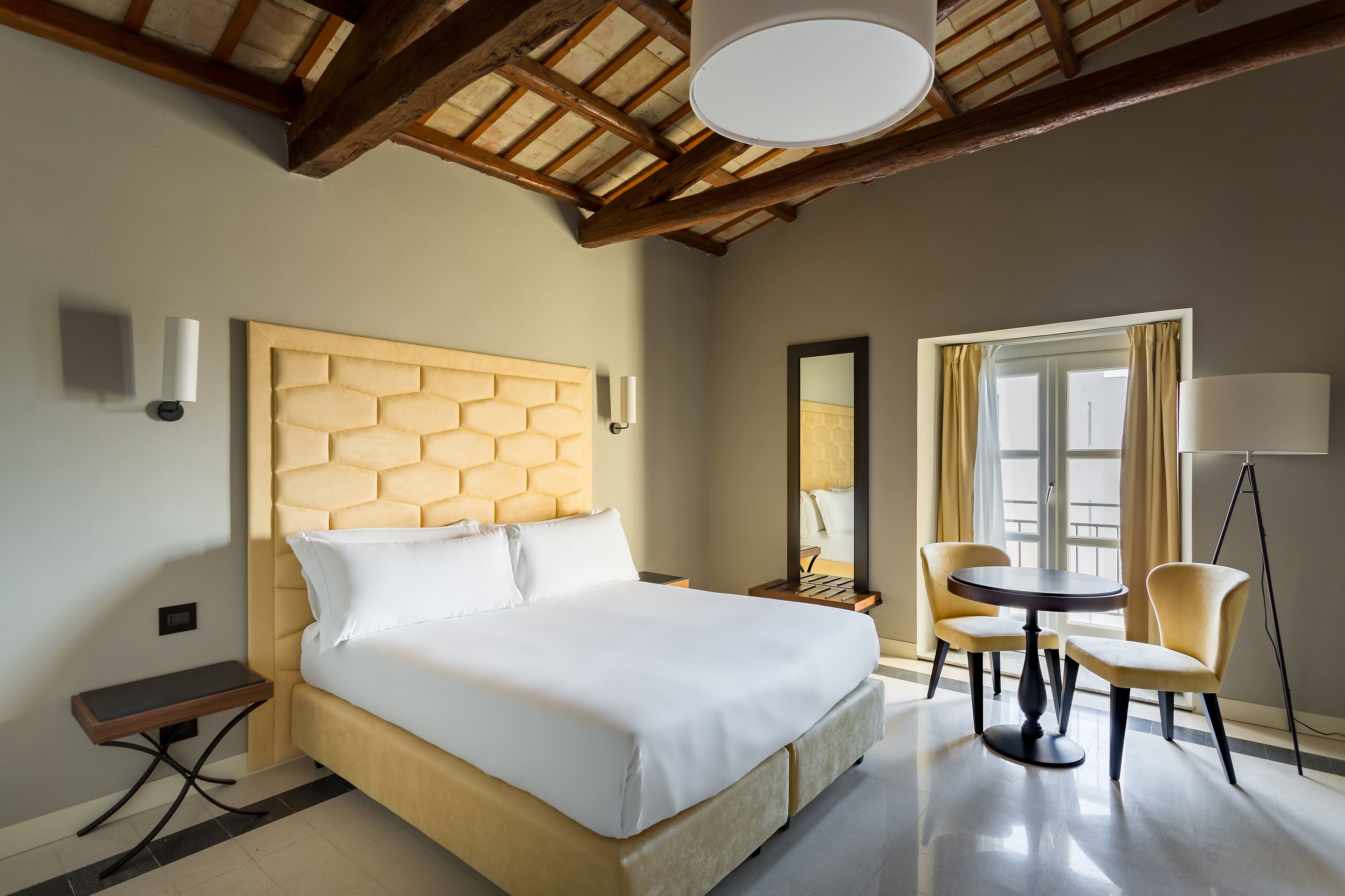 Ein Hotelzimmer mit einem großen Bett, einem runden Tisch, zwei Stühlen, einer Stehlampe und einem fensterbepflanzten Fenster.