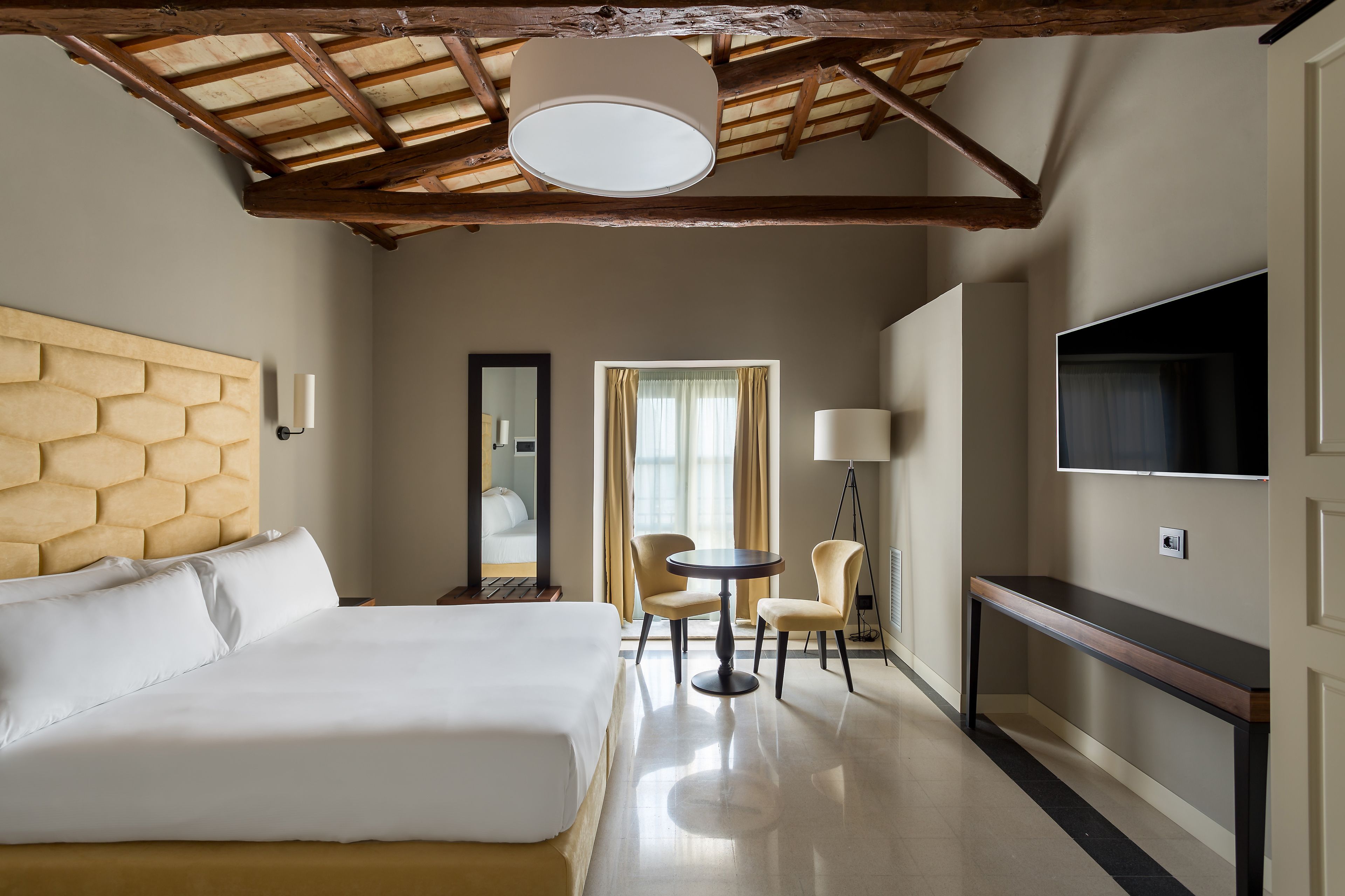 A hotel room with a large bed, a small round table, two chairs, a TV, and a mirror.