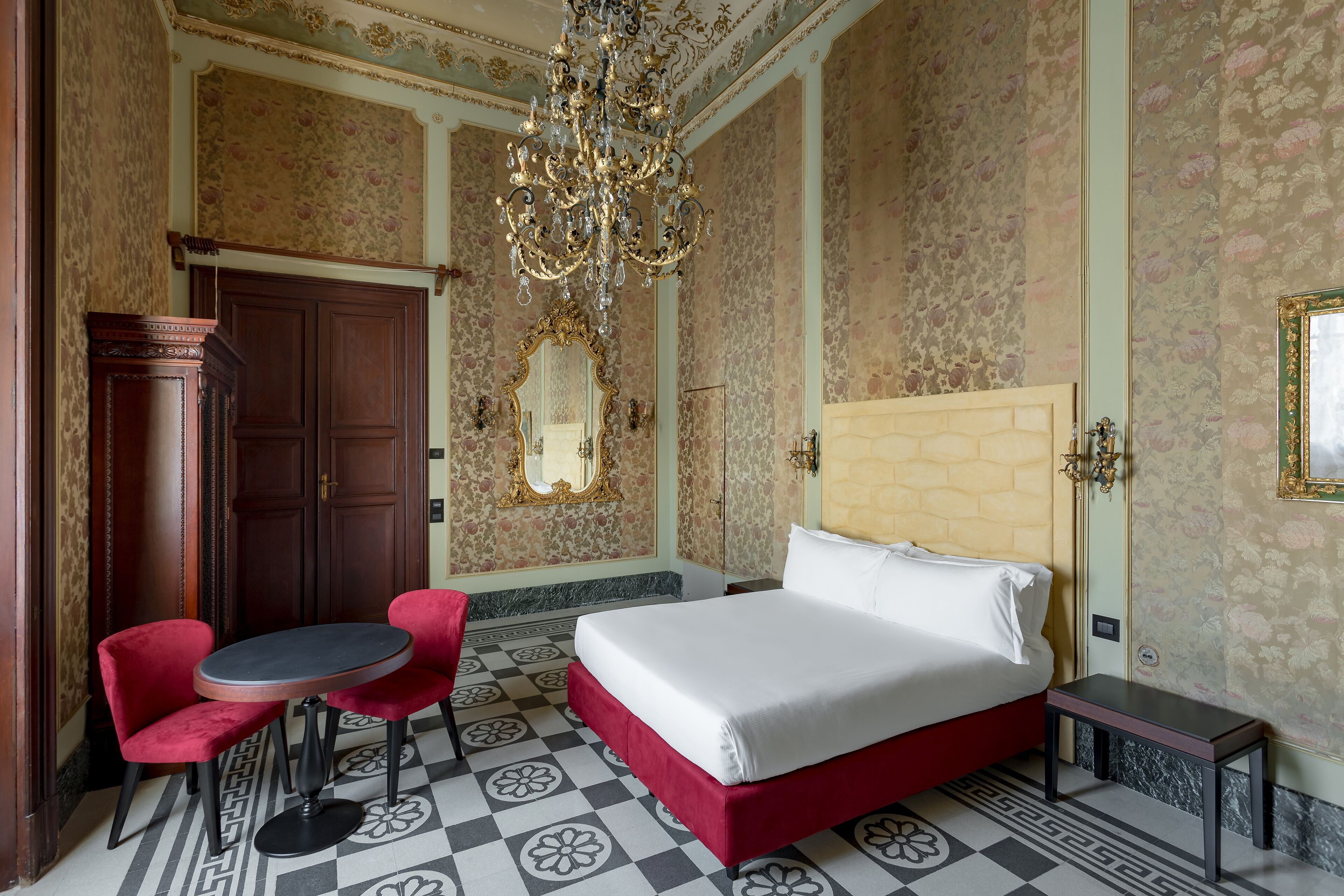 A hotel room with a bed, a round table with two chairs, a chandelier, and a large mirror.