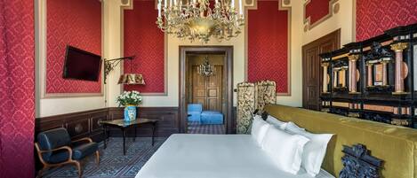 A luxurious hotel room with a large bed, a chandelier, a television, and a piano.