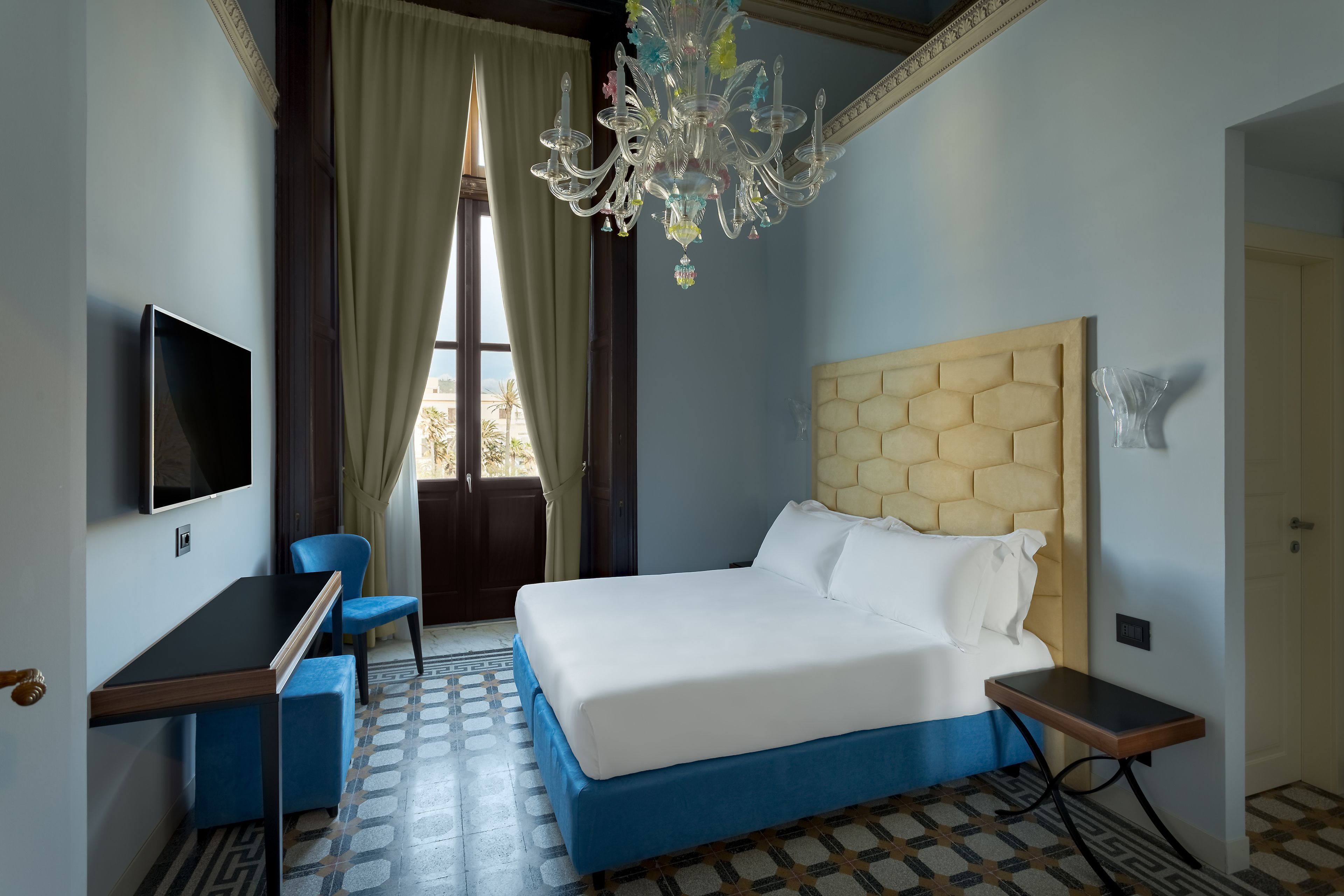 A hotel room with a bed, a desk, a chair, a television, and a chandelier.