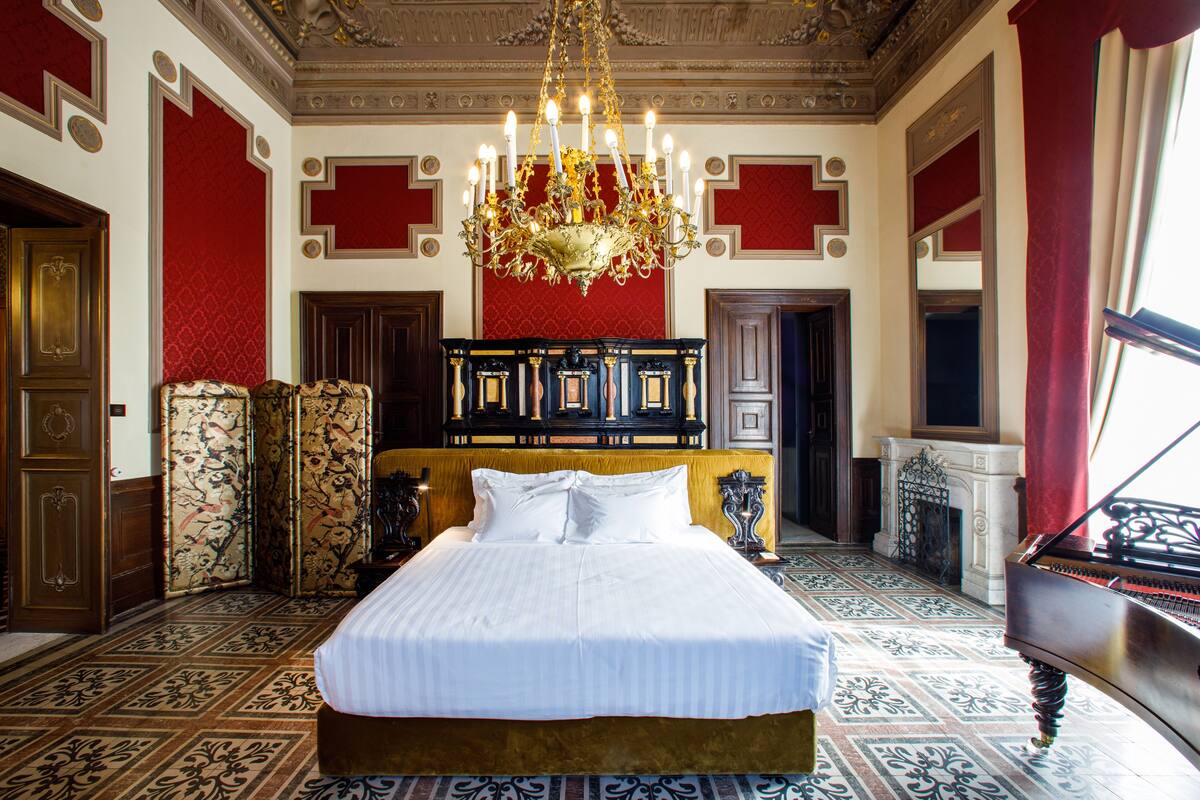 A large bed with a gold headboard, a chandelier, and a grand piano in a room with red and white walls and patterned flooring.