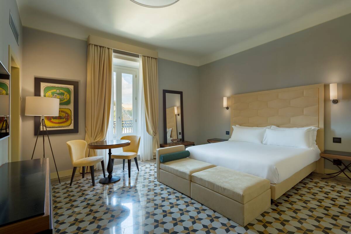 A hotel room with a large bed, a sofa, a small table, and a chair. There is a window with curtains, a framed picture on the wall, and a lamp.
