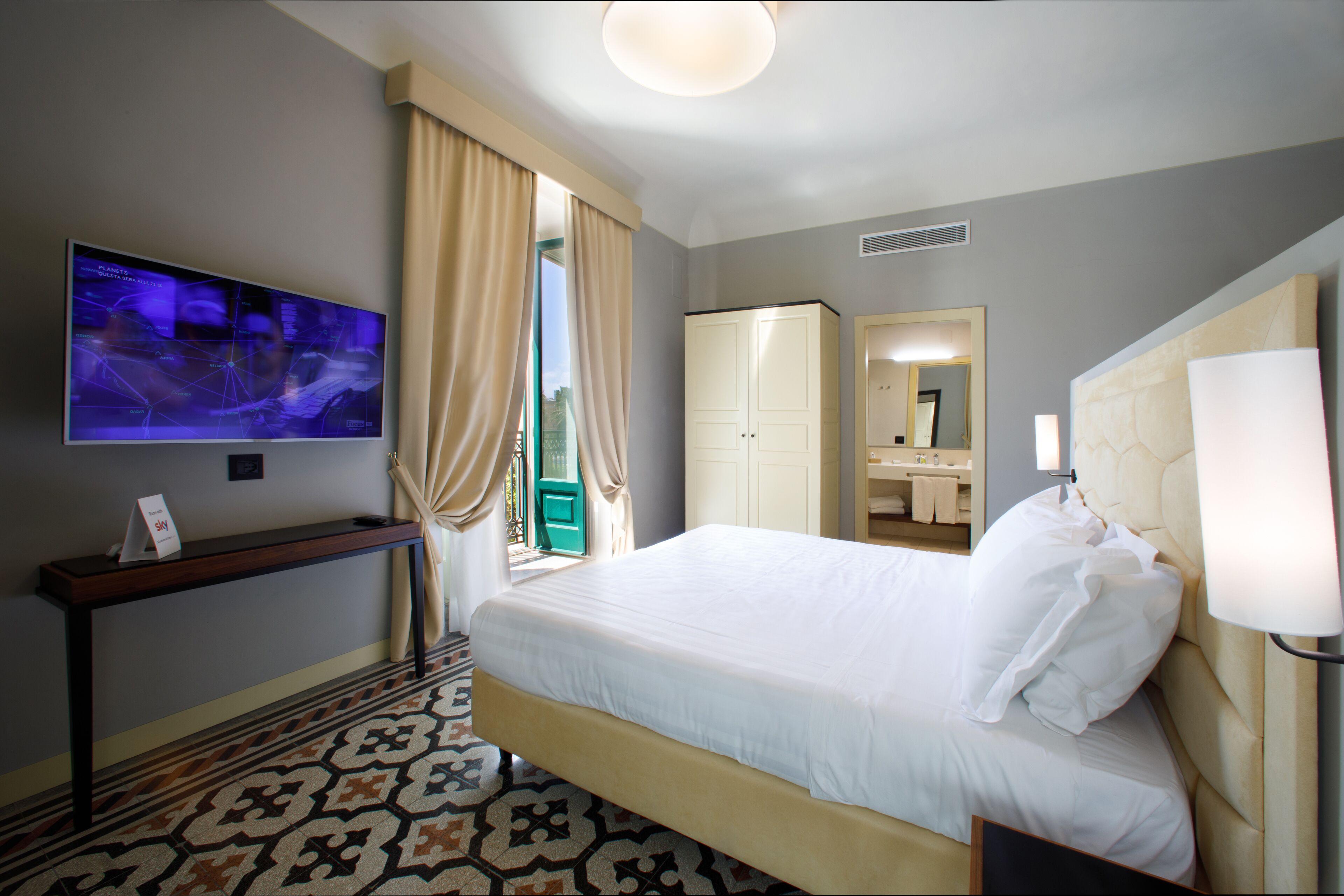 A hotel room with a bed, a TV, a desk, a wardrobe, and a bathroom visible through an open door.