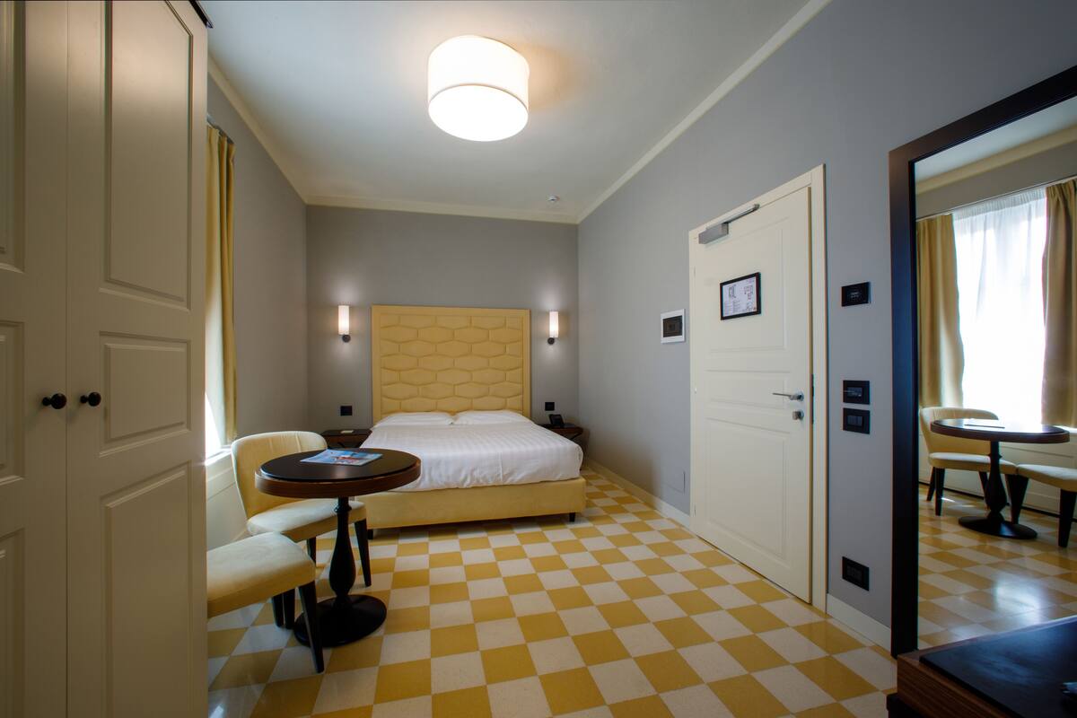 A hotel room with a checkered floor, a bed with a headboard, a bedside table, a chair, and a small table.
